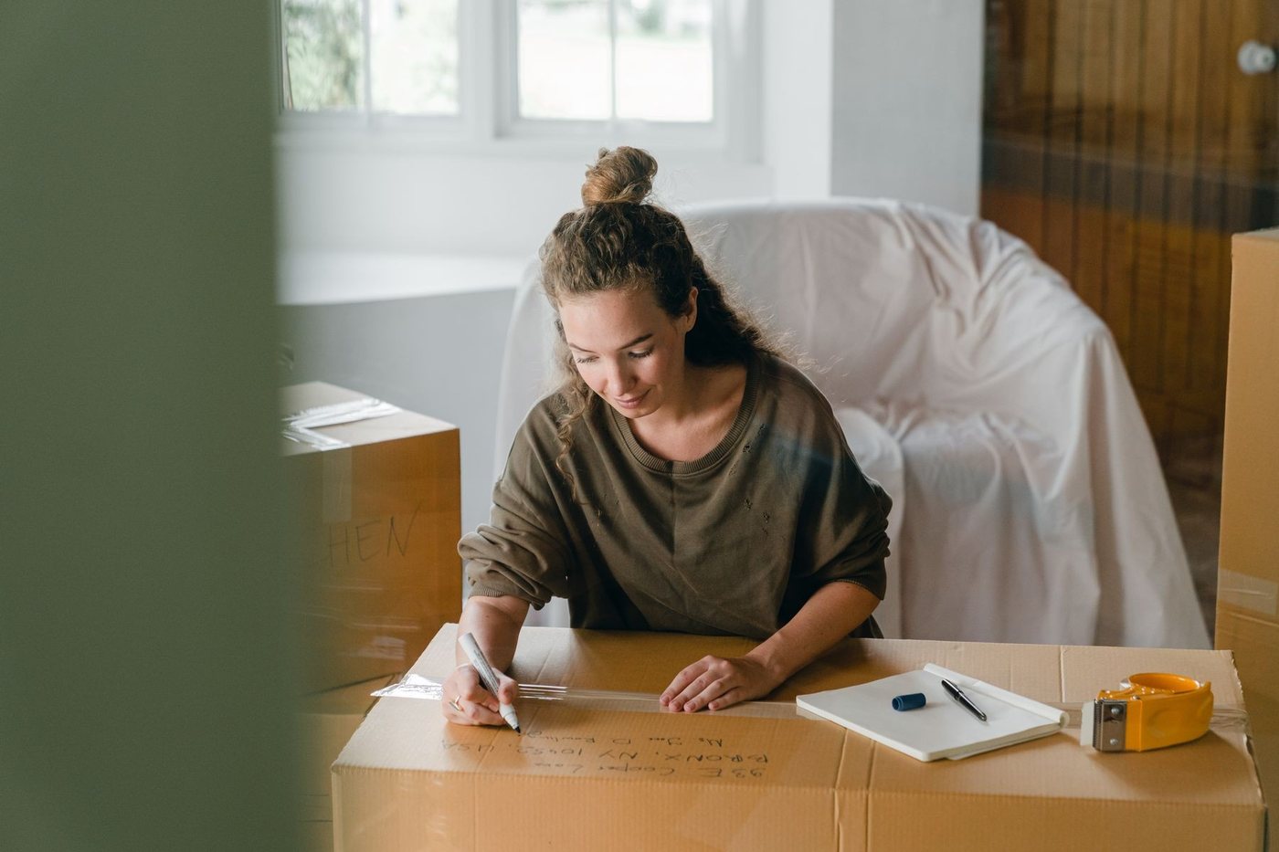 A woman labelling a packed carton in a half-cleared room, dust sheets over the furniture.